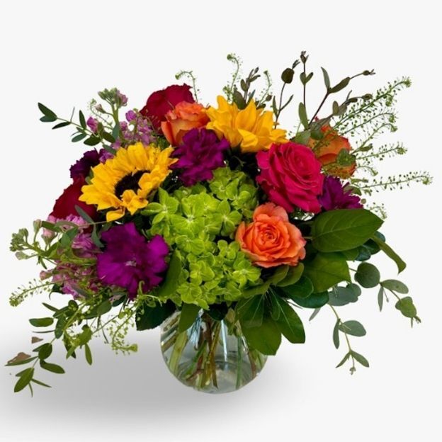 Bright bouquet with sunflowers, hot pink and orange roses, purple blooms, and green hydrangea in a round glass vase.