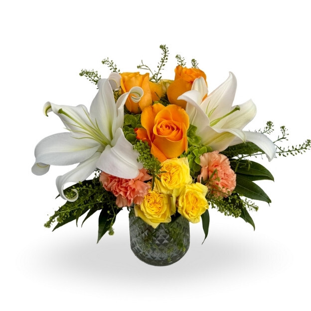 Bright floral arrangement featuring white lilies, orange and yellow roses, and mixed seasonal blooms in a textured glass vase.